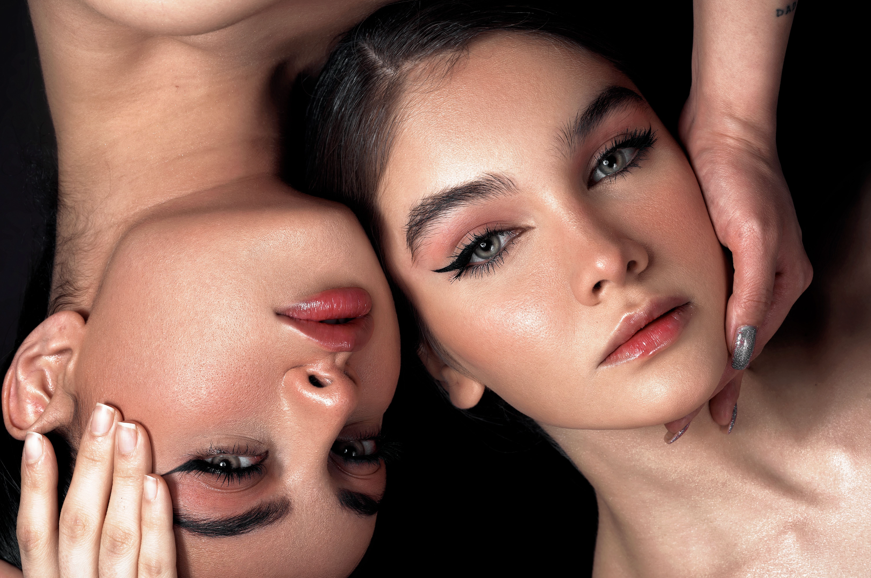 Close-up beauty shot of two women with dramatic eye makeup, glossy lips, and styled hair against a dark background, with a hand resting on one face.